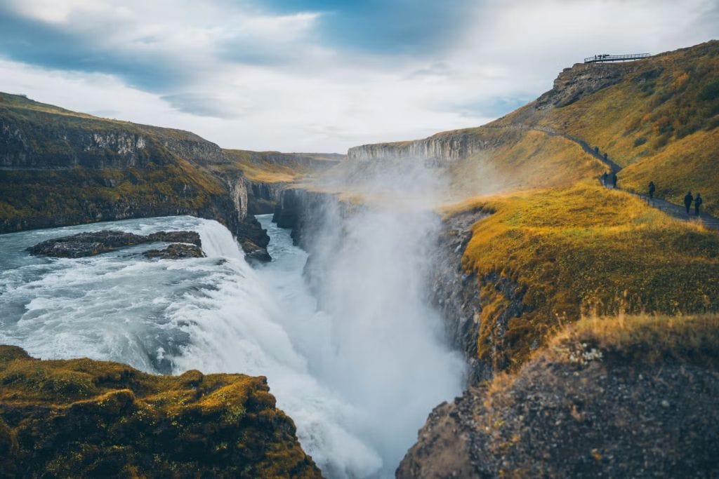 A Gullfoss vízesés Izlandon, ahol a hatalmas víztömeg két lépcsőben zuhan a mély kanyonba. A környező zöld és aranyszínű domboldalakon túrázók sétálnak a kilátópont felé.