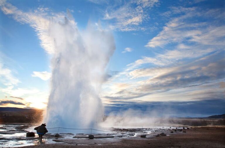 A Strokkur gejzír kitörése a Geysir geotermikus területen Izlandon, a naplemente fényében. Egy turista figyeli a magasba törő vízoszlopot a gőzölgő földfelszín mellett.