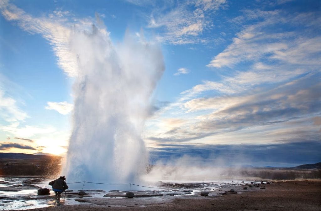 A Strokkur gejzír kitörése a Geysir geotermikus területen Izlandon, a naplemente fényében. Egy turista figyeli a magasba törő vízoszlopot a gőzölgő földfelszín mellett.