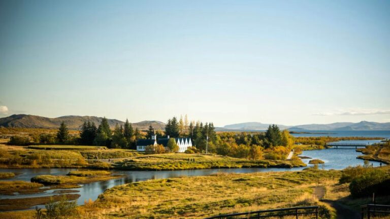 A Þingvellir Nemzeti Park napsütéses tája Izlandon, előtérben folyókkal és zöldellő rétekkel, háttérben a fehér Þingvallakirkja templom és a környező hegyek láthatók.
