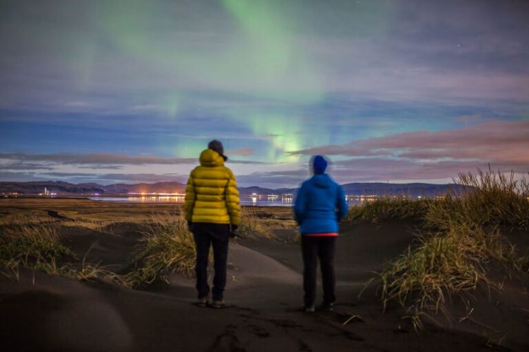 Két ember áll a sötét homokdűnéken, és a város fényei fölött táncoló sarki fényt nézik Izlandon.