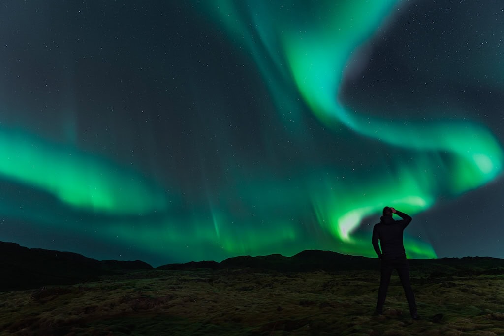 Egy ember sziluettje ahogyan a zölden táncoló sarki fényt kémleli.