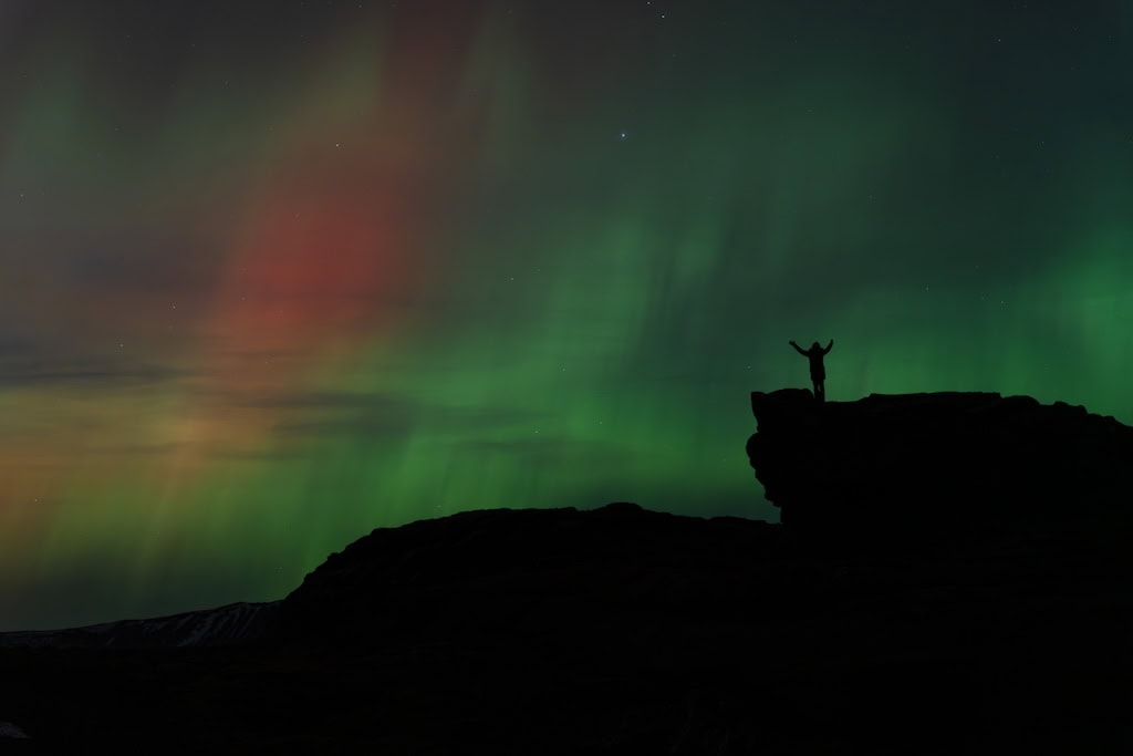 Sarki fény Izland éjszakai égboltján halvány vörös és zöld fátyolként, sötét sziklák körvonalával.