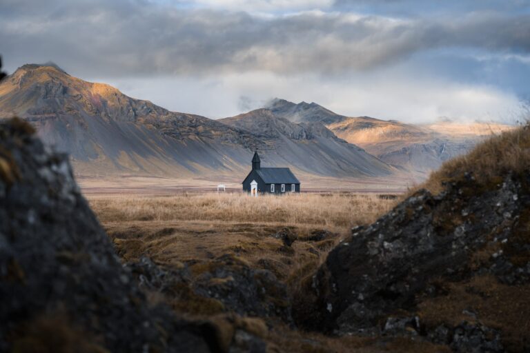 A híres fekete fatemplom Búðirban, a Snæfellsnes-félszigeten, háttérben a napfényben megvilágított hegyekkel és drámai felhőkkel.
