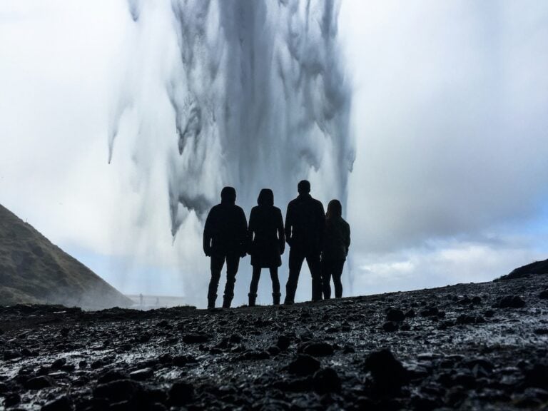 Utazók sziluettje a Seljalandsfoss-vízesés mögött Izlandon, fekete kavicsos parttal és vízpermettel.